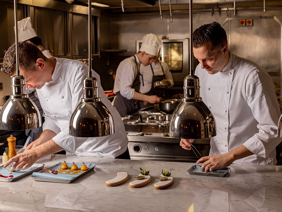 Chefs in white uniforms meticulously plating gourmet dishes under heat lamps in a professional kitchen, with two chefs in the foreground focusing on presentation and two more cooking in the background.