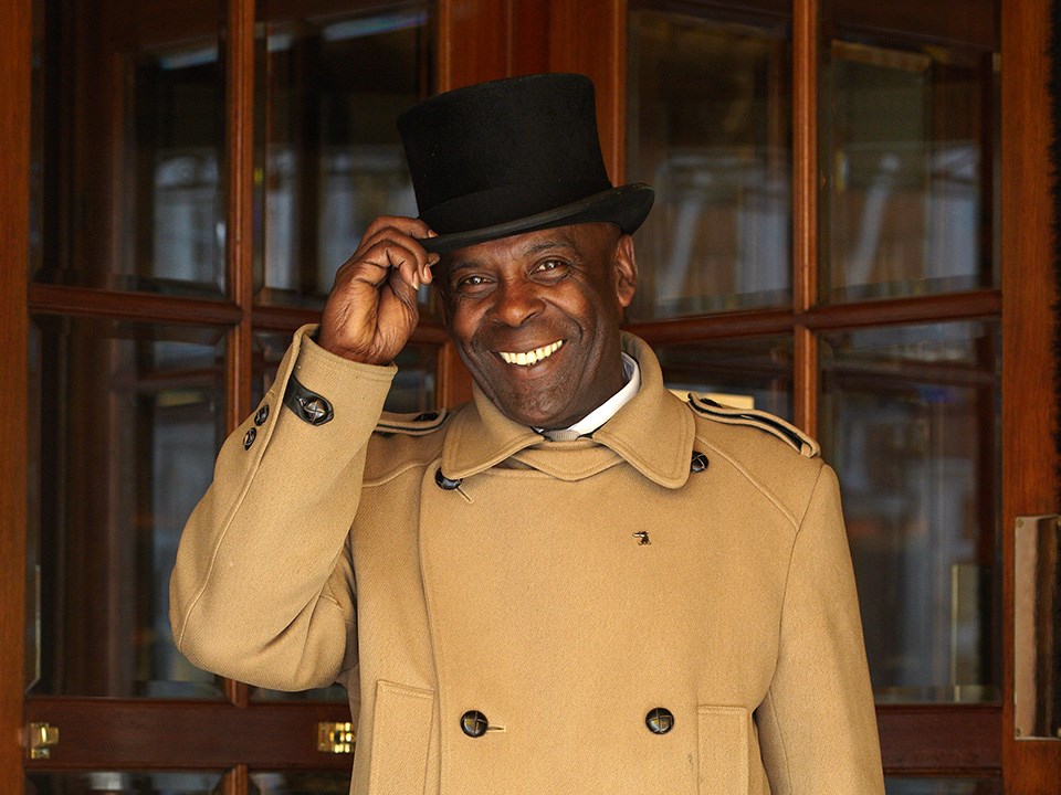 Portrait of a smiling doorman with a hat in front of a hotel, greeting his guests.