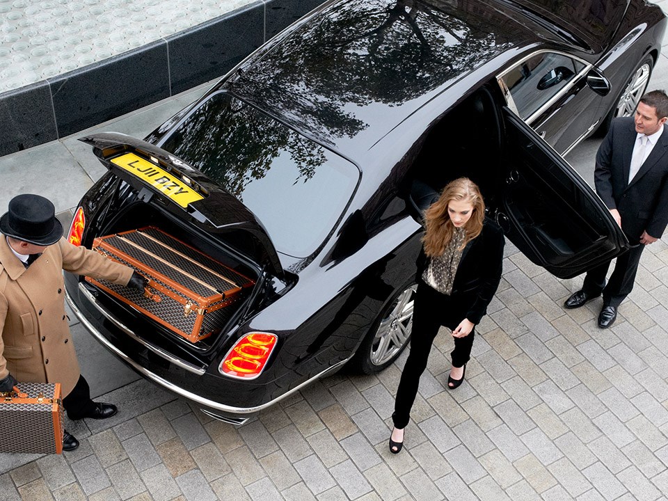 A concierge taking travel bags out of a car, and an incoming hotel guest stepping from the car.