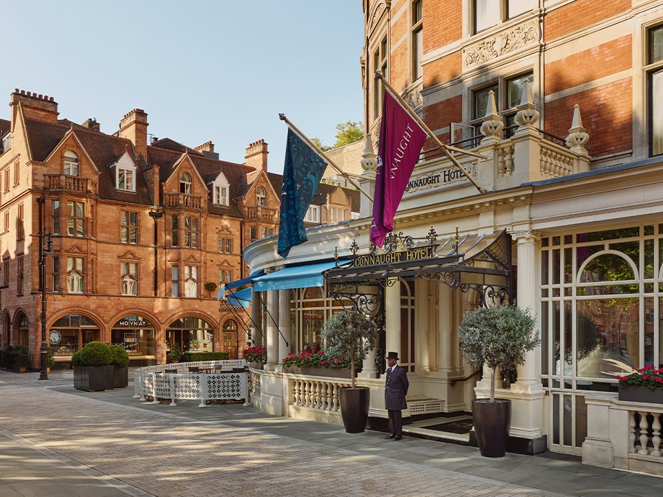 Exterior view of The Connaught Hotel with flags, a doorman at the entrance, and surrounding historic buildings in daylight.