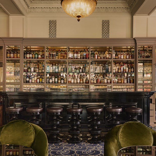 Refined interior of the Coburg Bar, featuring a dark marble bar with leather stools, an extensive backlit display of premium spirits, and a decorative ceiling with a central chandelier.