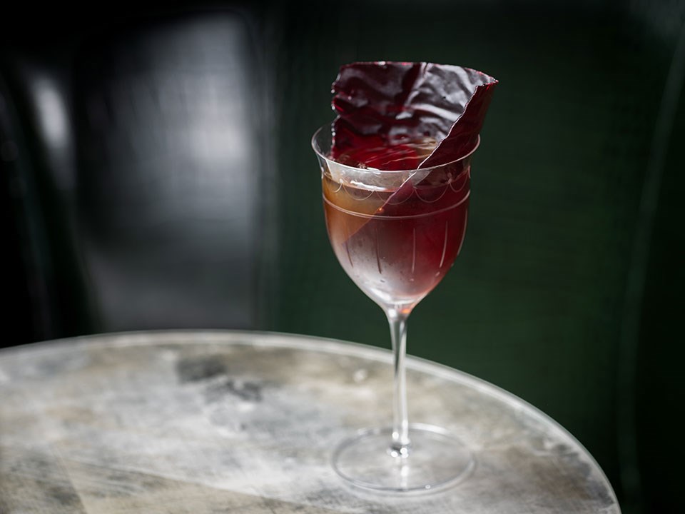 connaught bar burgundy cocktail in tall thin stemmed glass on a glass table with a dark green background
