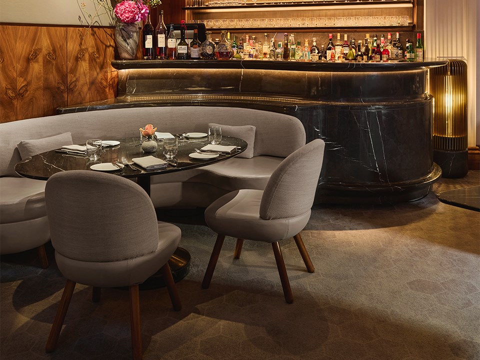 An elegant dining setup with a curved banquette and plush grey chairs surrounding a dark marble table, set for service, with a softly lit bar in the background showcasing premium spirits and floral accents.
