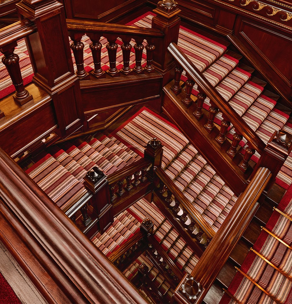 Ornate wooden staircase with striped red and beige carpet runners, viewed from above in a multi-level interior.