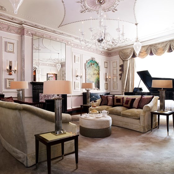 The living room at the Sutherland Suite. Two comfortable sofas face a central pouffe which has cake and and tea on it. Behind them, there is a grand piano in front of a large window. On the walls there are paintings of birds and a large mirror.