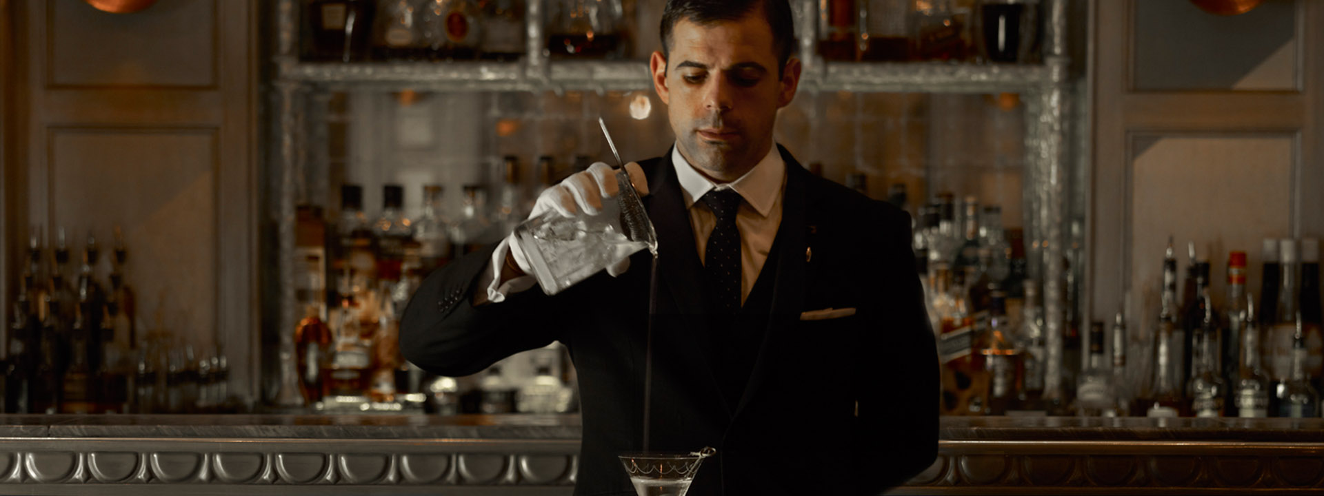 A bartender dressed in a suit, pouring drink from a vessle to a glass with a dim light.
