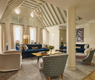 Elegant lounge with high vaulted ceiling, tall windows with light curtains, blue velvet sofas, and patterned armchairs around a central coffee table.