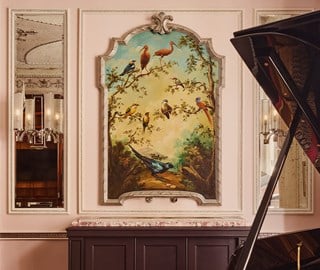 Classical interior with a large framed painting of colorful birds on a tree, flanked by mirrors and a grand piano in the foreground.