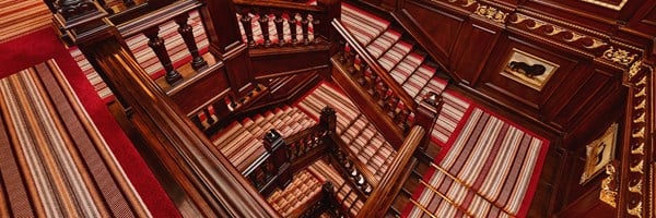 Wide-angle view of a richly detailed wooden staircase with red and beige striped carpeting, gold trim, and framed artwork on the walls.