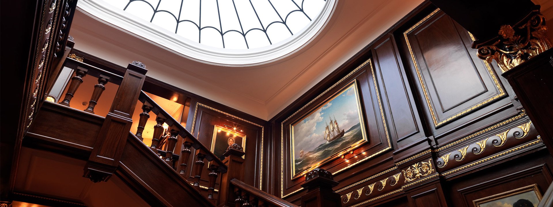 The staircase at The Connaught featuring paintings with golden frames hanged on wooden walls.