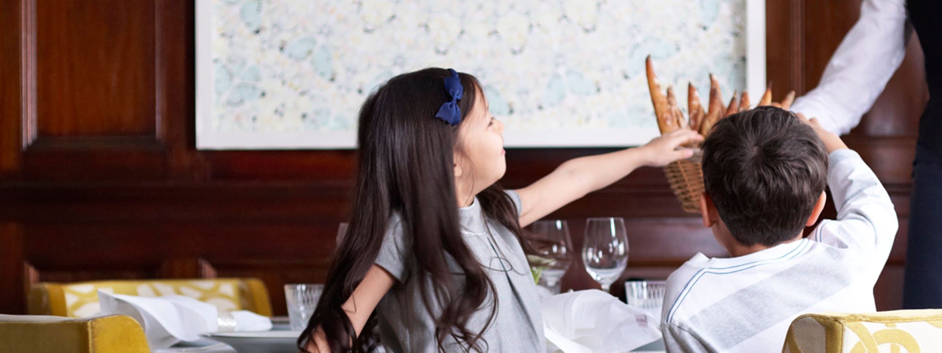 Two children sitting in a restaurant taking food from a waiter's tray.