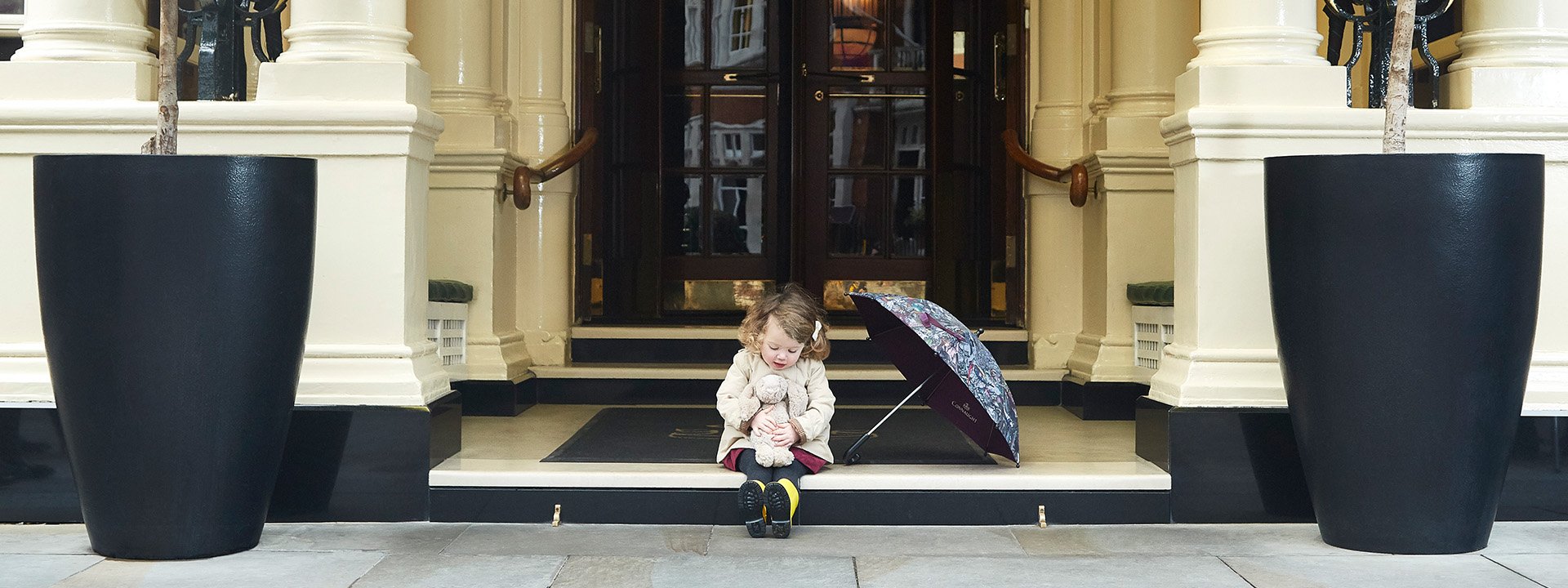 
A little girl with a stuffed bunny next to an open umbrella, sitting at the entrance of The Connaught hotel.