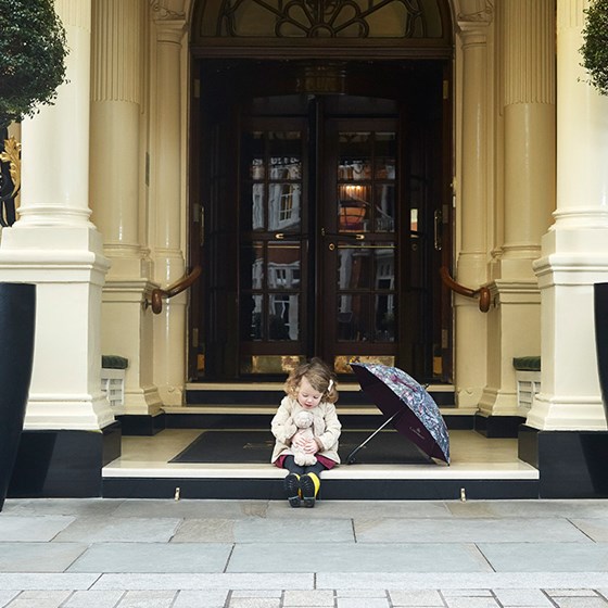 Photo of a playful little girl in front of the entrance, playing with her teddy bear.