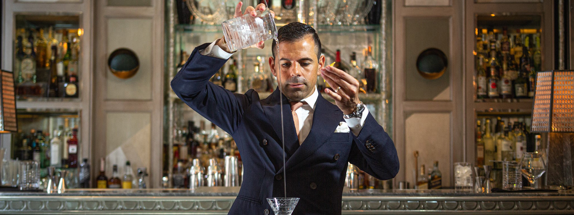 A bartender dressed in a suit, pouring a drink from a vessel to a glass.