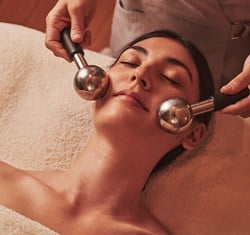 Spa therapist performing facial massage using metal globes on a relaxed woman lying on a treatment bed with closed eyes.