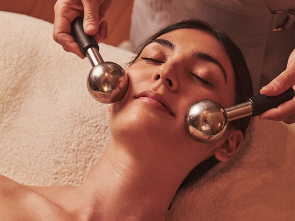 Woman receiving facial massage with metal globes, lying on a treatment bed with eyes closed in a spa setting.