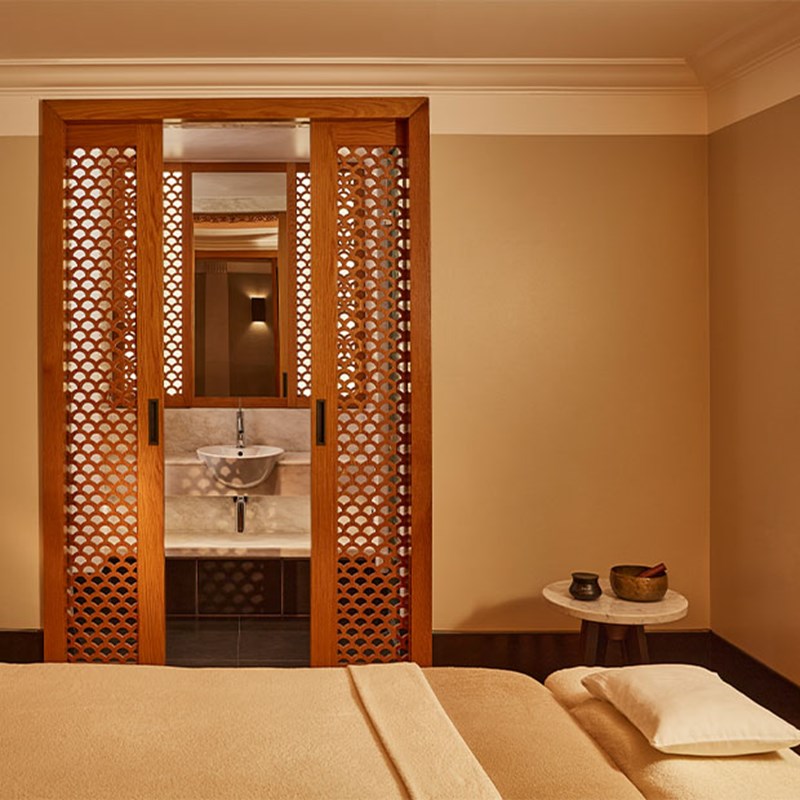 Alt text: Luxury spa room with a central massage bed, warm lighting, wooden lattice screens, and a marble washbasin behind.