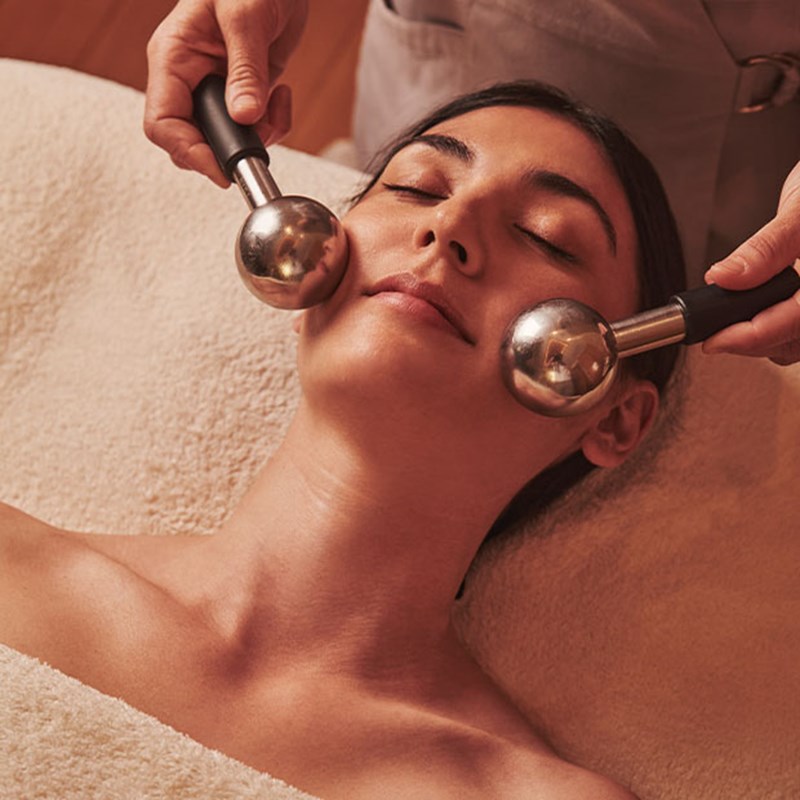 Spa therapist using metal facial rollers during a treatment on a relaxed woman lying on a soft cream towel.