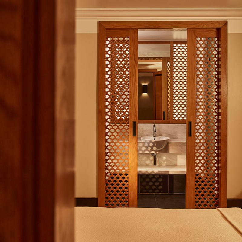 Elegant spa treatment room with a beige massage bed, warm lighting, and carved wooden screen framing a basin.