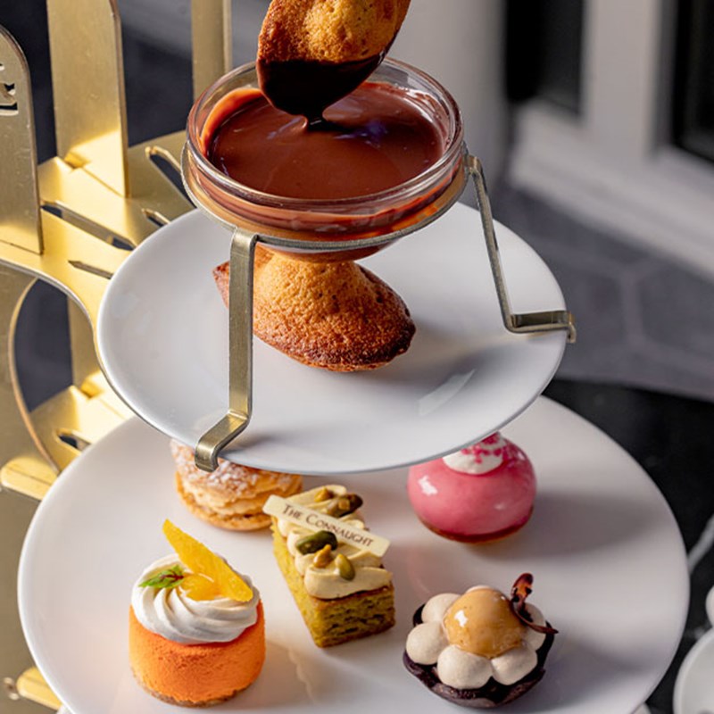 A refined afternoon tea presentation with a golden stand, featuring delicate madeleines dipped in rich chocolate sauce, complemented by a selection of exquisite pastries.