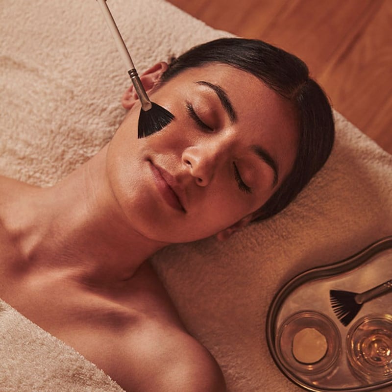 A tranquil spa scene where a woman enjoys a luxurious facial treatment, as a soft brush gently applies a rejuvenating mask in a warm, candlelit ambiance.
