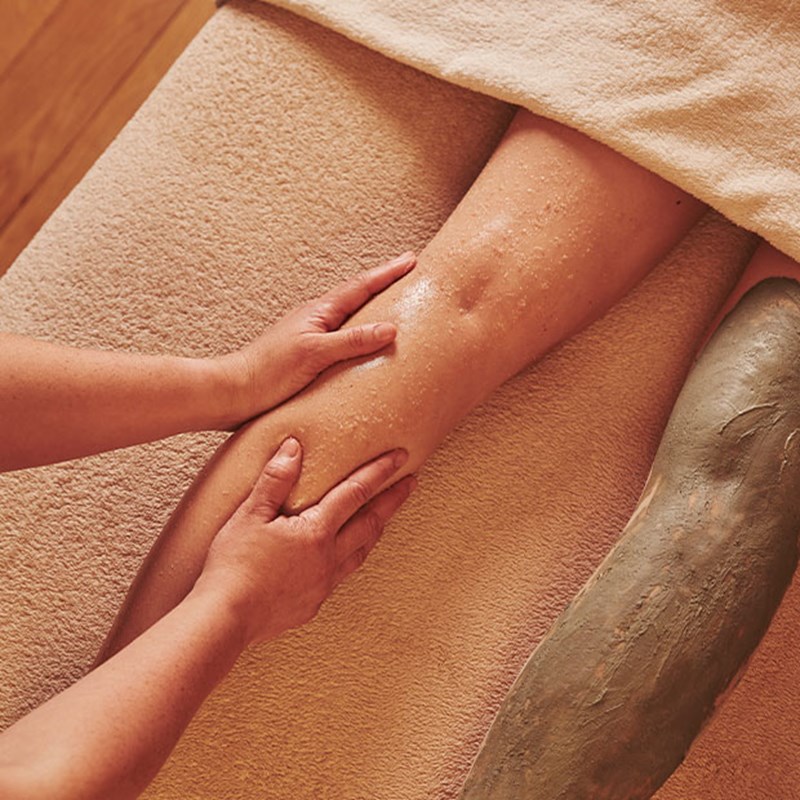 A serene wellness ritual in progress, with an expert therapist massaging an exfoliating treatment into a guest’s leg, accompanied by a soothing body wrap.