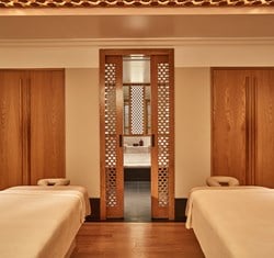 A tranquil spa treatment room with two plush massage beds, warm wooden accents, and softly lit lattice-panel sliding doors leading to a serene wash area, evoking a sense of calm and indulgence.