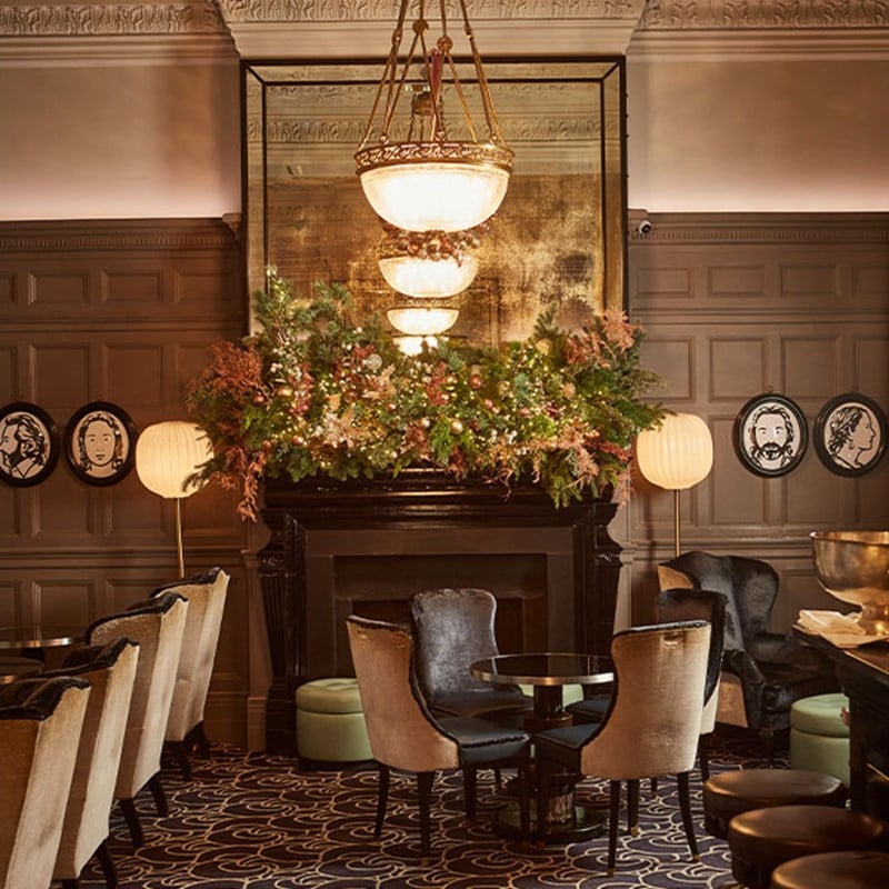 A cosy room with round tables and chairs, and a mantelpiece decorated with festive, light up greenery.
