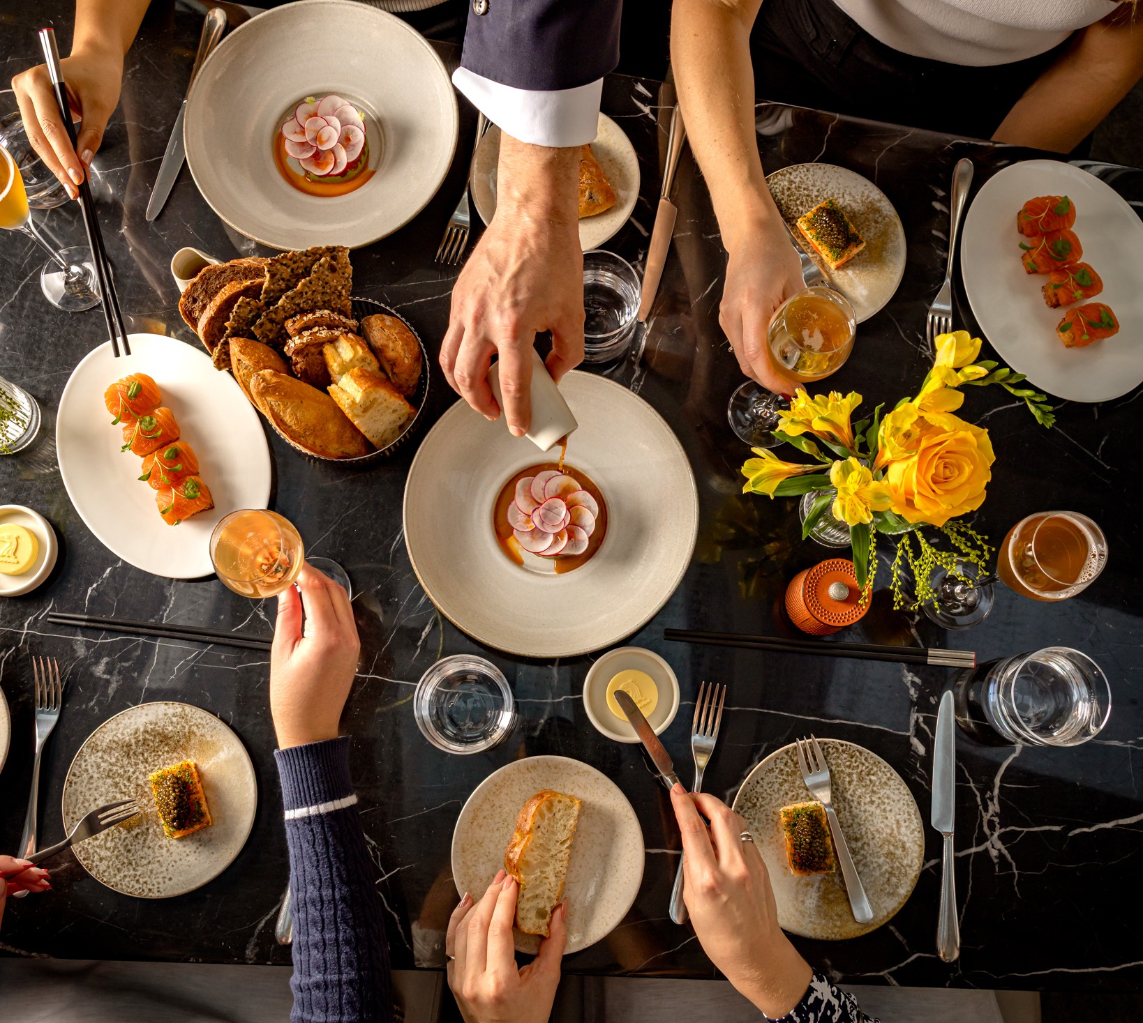 Overhead view of a shared fine-dining table with plated dishes, bread and drinks as diners reach in to serve and eat.