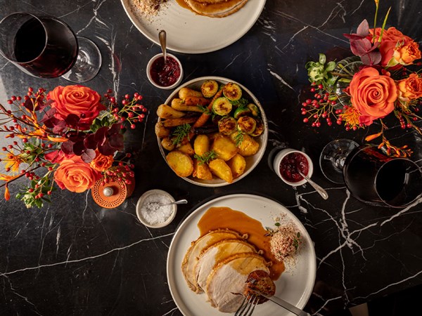Overhead view of two diners cutting roast turkey with gravy, sides and wine on a dark table decorated with bright floral arrangements.