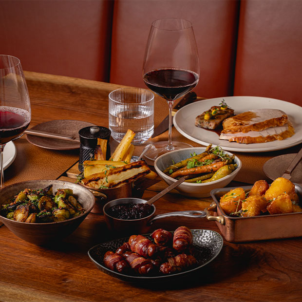 A festive table spread with roast turkey slices, vegetables, roast potatoes, pigs in blankets, red wine, and assorted sauces in small bowls.