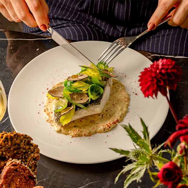A plated main course of white fish topped with truffle slices and green leaves, served on a creamy sauce as a person cuts into it with a knife and fork.