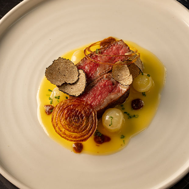 A gourmet meat dish featuring a seared cut of beef with truffle slices, caramelised onion, pearl onions, and a golden sauce elegantly plated.