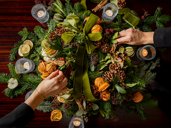 Hands adjust a festive wreath of evergreens, dried citrus, pinecones and a green velvet bow on a painted door