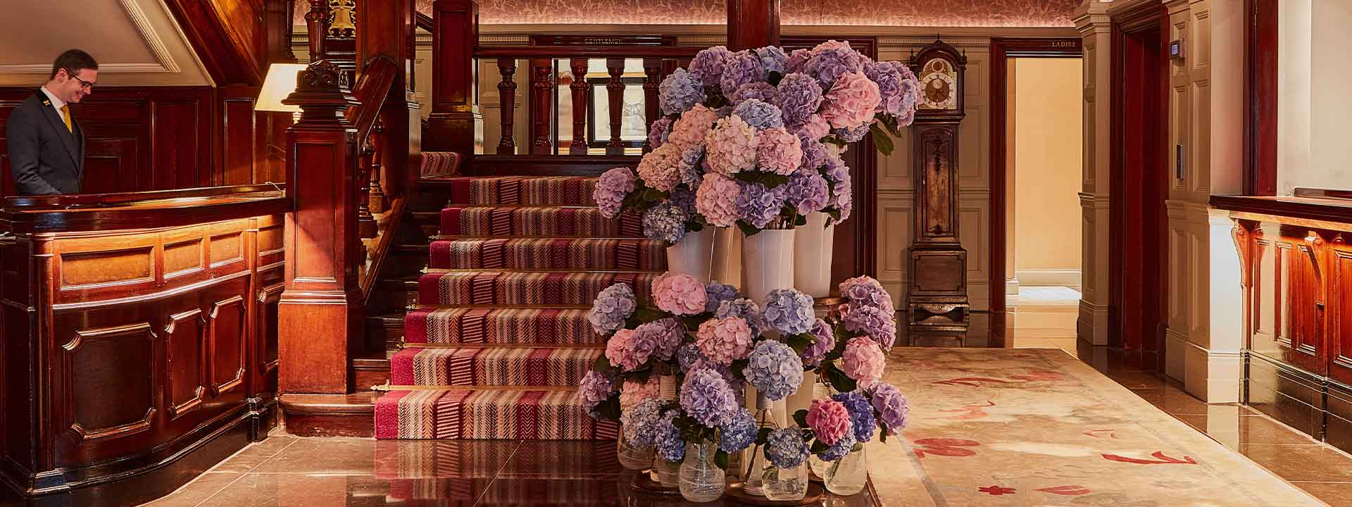 A hotel concierge is working smilingly at The Connaught, next to magnificent flower arrangements.