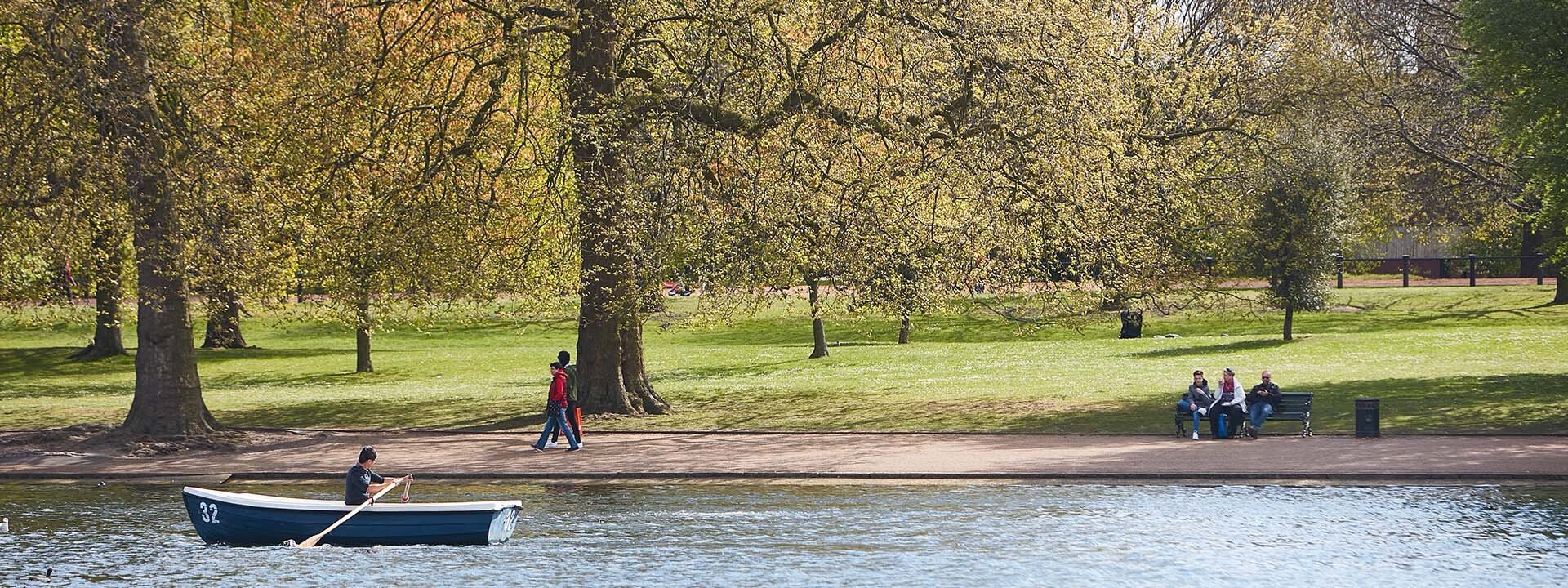 Trees and a lake with a little boat being rowed by a person in Hyde Park.