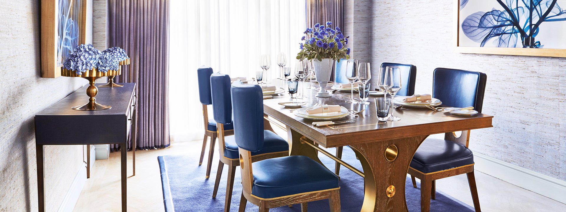 Long wooden table, with glasses and plates, with six dark blue chairs, in a room with natural light.