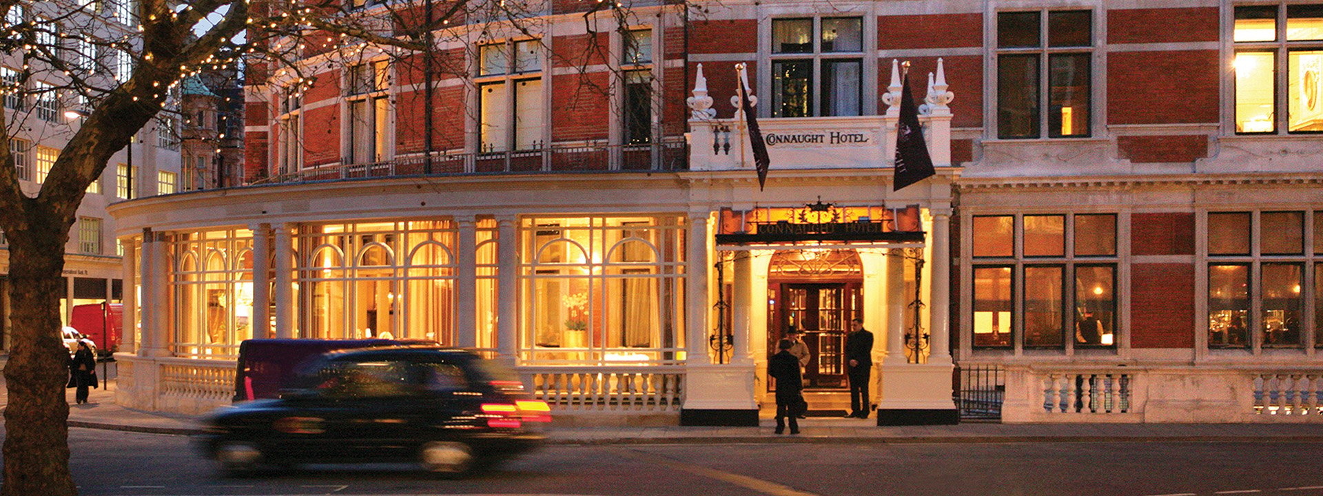 View of The Connaught, from the outside, with a car passing and concierges in front.