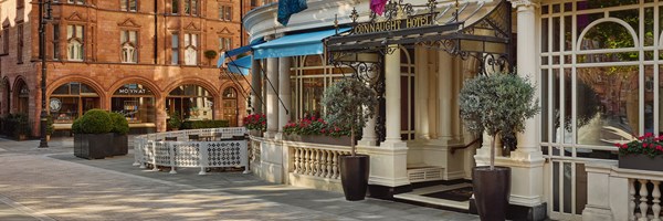 Close-up of The Connaught Hotel entrance with ornate canopy, potted trees, flower boxes, and outdoor seating area beside a red brick building.