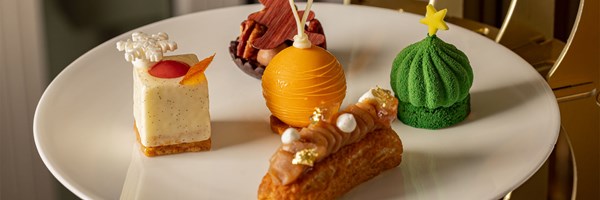 Assorted festive pastries on a white plate: vanilla cube, orange sphere, green dome and chestnut éclair.