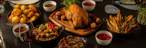 Festive roast dinner featuring golden turkey, roasted potatoes, glazed carrots, Brussels sprouts, and cranberry sauce on a dark table.