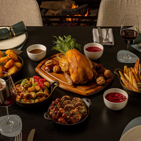 Festive roast dinner set before a fireplace, featuring a golden roasted turkey, vegetables, potatoes, cranberry sauce, and red wine on a dark dining table.