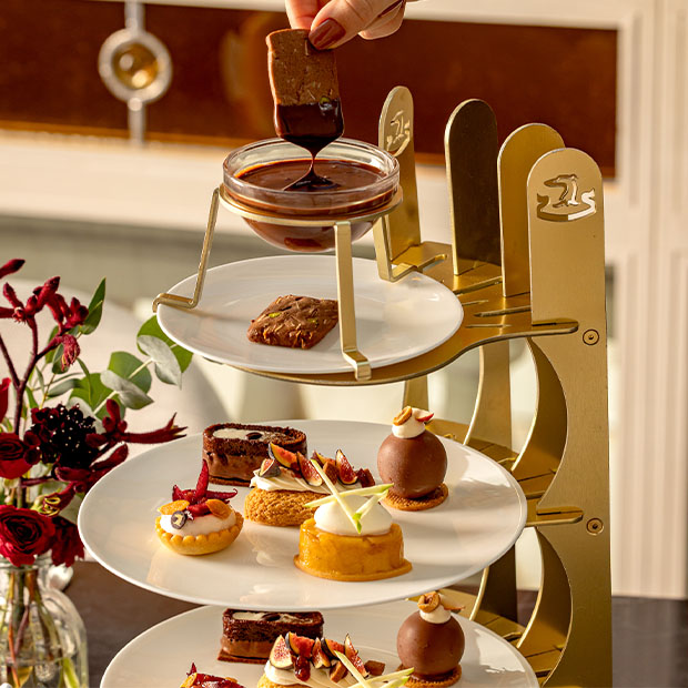 Hand dipping a biscuit into warm chocolate on a gold-tiered stand displaying an assortment of elegant pastries and desserts.