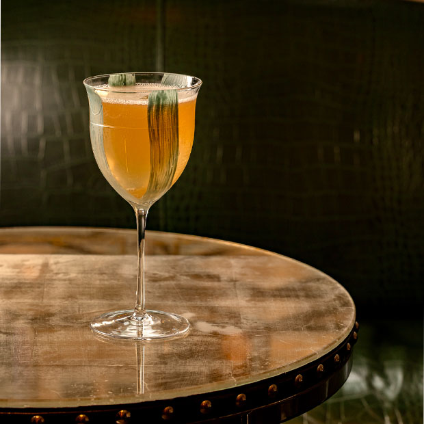 Amber cocktail in a stemmed glass garnished with a green leaf, placed on a round marble-topped table.