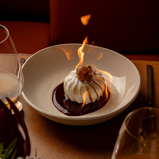 Baked Alaska dessert set alight at the table, served in a white bowl with rich chocolate sauce.