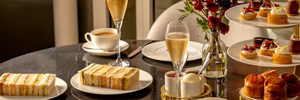 Afternoon tea setting with finger sandwiches, scones, pastries, champagne, and coffee arranged on a black marble table with a vase of red flowers.