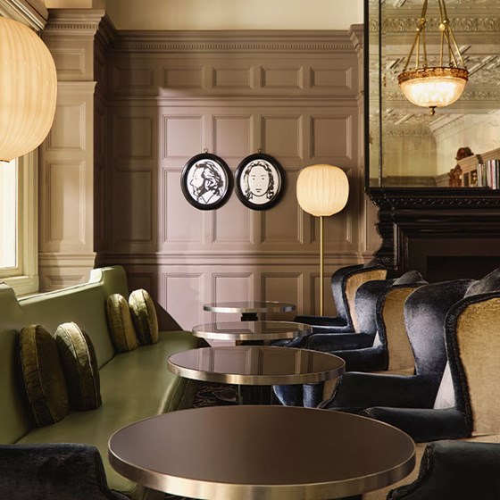 A refined lounge setting with paneled walls, velvet armchairs, and olive-toned banquette seating, accented by soft globe lighting and monochrome portrait art for a touch of modern whimsy.