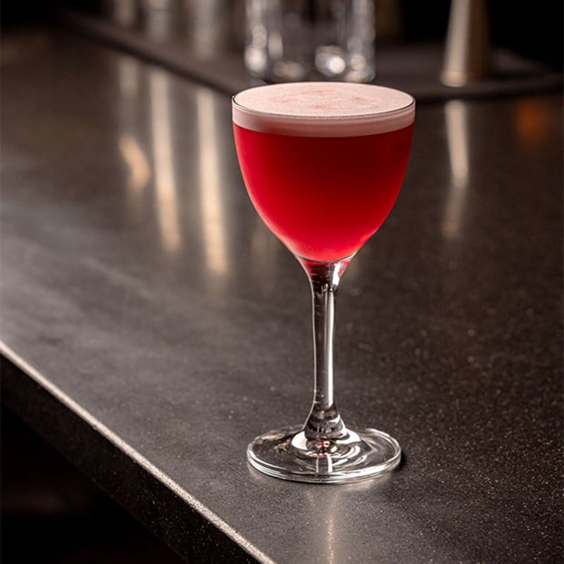 A vibrant pink cocktail with a foamy top served in a stemmed glass on a dark bar counter.