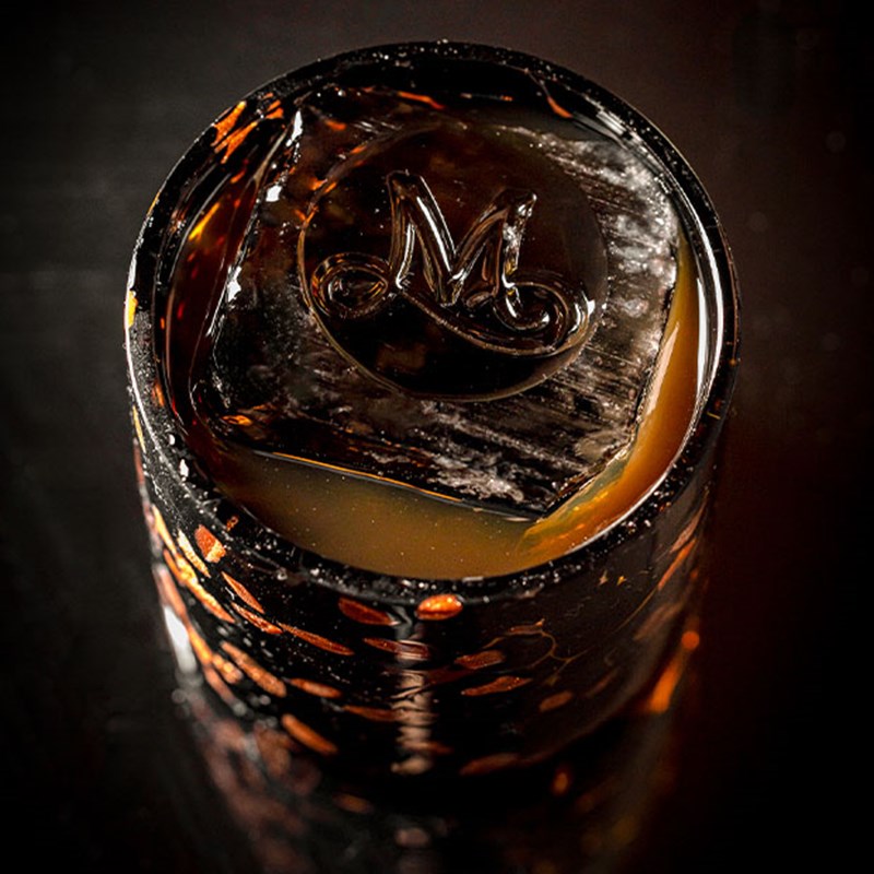 A rich, dark cocktail in a patterned glass with a large ice cube engraved with a cursive “M”.