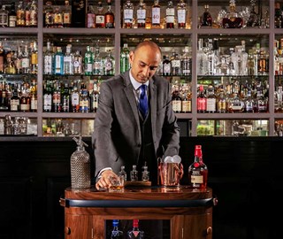 A bartender focused on preparing whiskey drinks, with a warm atmosphere against the background of a shelf with many drinks.